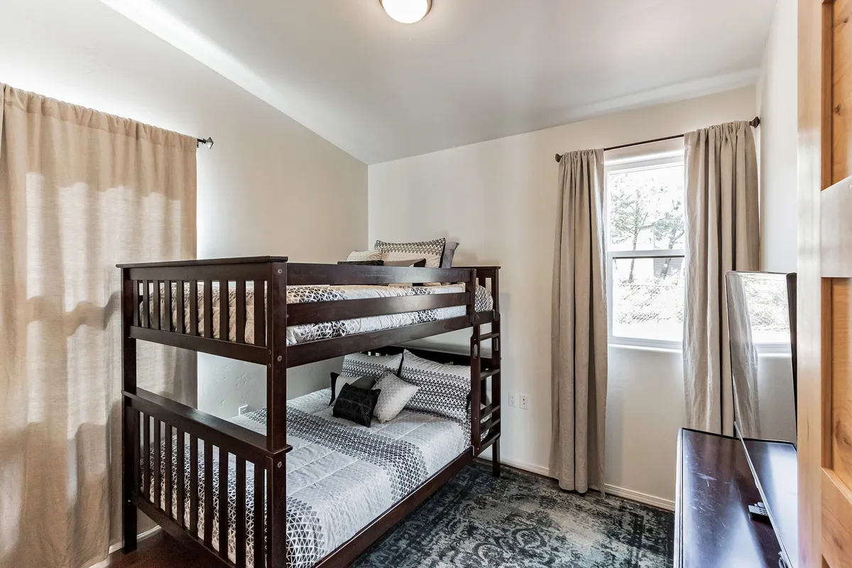 A cozy bunk bedroom with sturdy wooden bunk beds, playful decorative pillows, and a window with flowing curtains. The space is bright and inviting, ideal for children or extra guests, and evokes a sense of fun and togetherness.