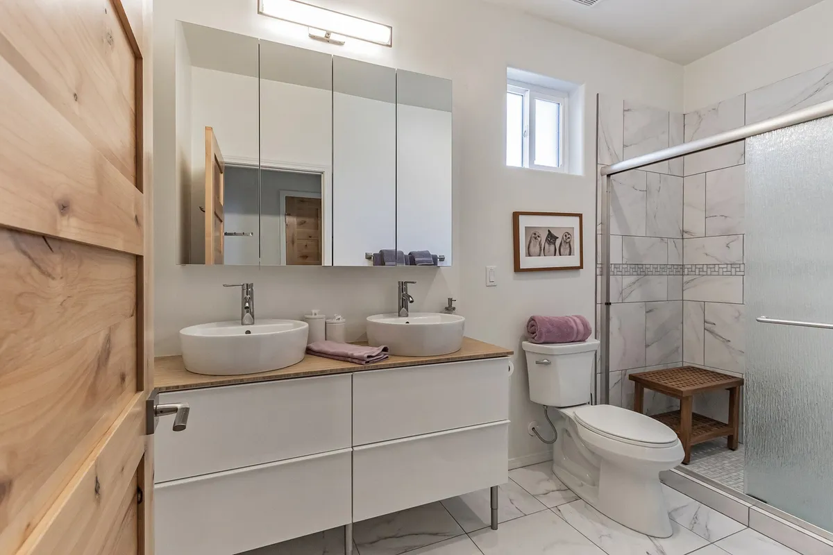 A modern bathroom featuring double vessel sinks, expansive mirrors, a glass-enclosed shower, and warm wooden accents. The space is bright, clean, and thoughtfully designed for self-care and relaxation.