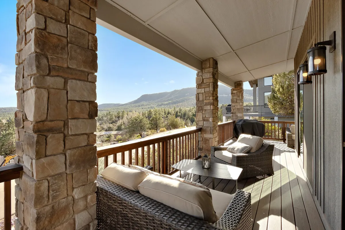 A spacious deck with sturdy stone pillars, comfortable outdoor seating, and a glass railing opens up to an expansive view of the mountains. The sun is low in the sky, suggesting the golden hour—perfect for sunsets or stargazing. The design seamlessly blurs the boundary between indoor and outdoor living, inviting relaxation and socializing against a mountain backdrop.