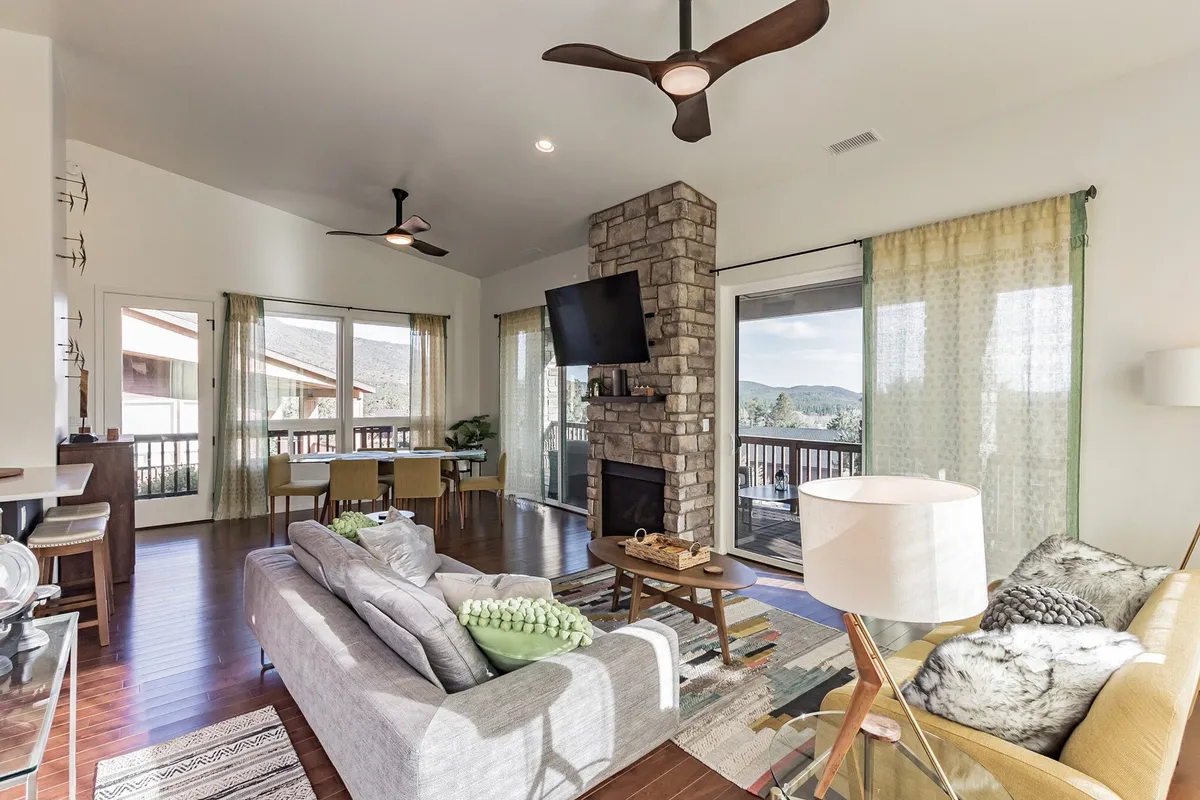 This image features a cozy great room with a fireplace, large windows showcasing mountain views, a comfortable sofa, and a spacious dining table. Natural light floods the room, enhancing the inviting atmosphere.