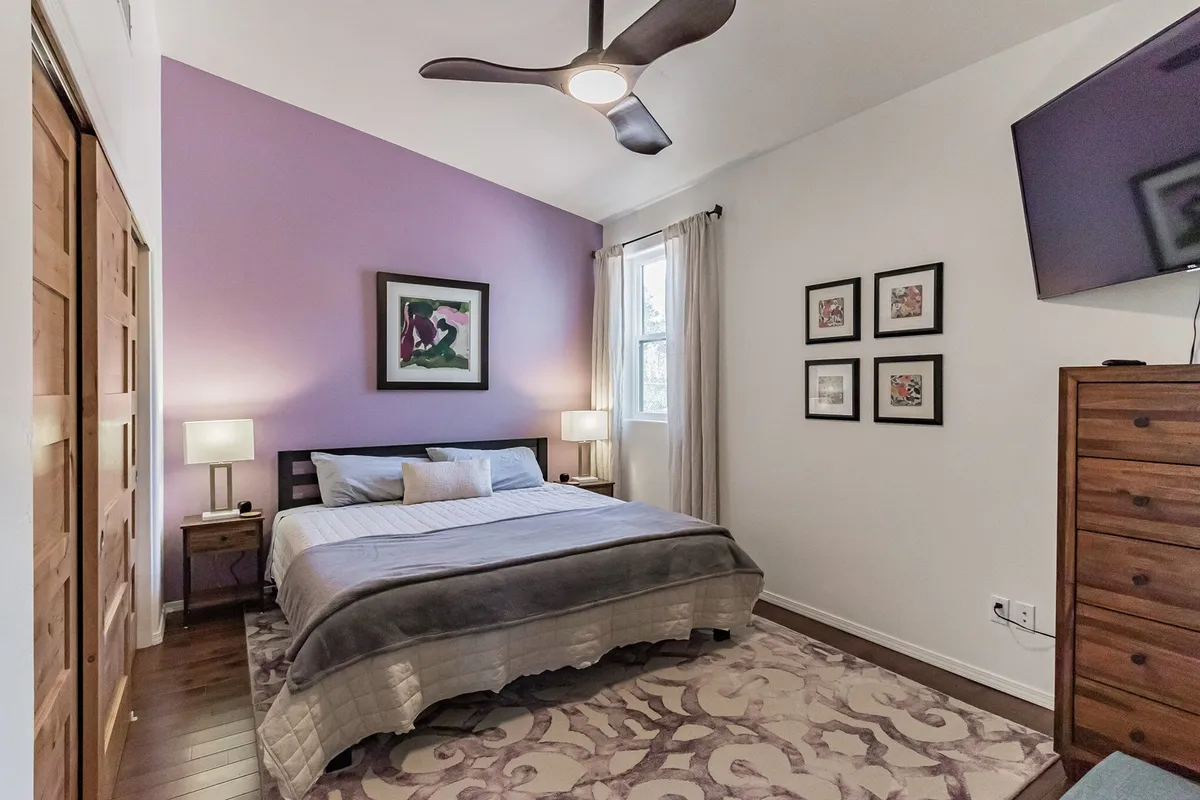 A tranquil bedroom featuring a neatly made bed with soft linens, a contemporary artwork above the headboard, a warm lamp on a bedside table, and a simple dresser. Sunlight filters into the space, adding a peaceful, inviting atmosphere ideal for restful sleep.